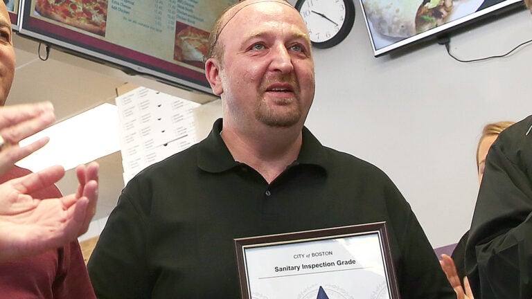 Stash's Pizza owner Stavros Papantoniadis is shown wearing a black collared shirt and holding up an "A" grade sanitary inspection certificate.