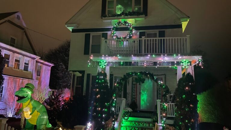 Chris and Daisy Hugenberger's Roslindale home, decorated with green and white holiday lights, a shamrock arch, and an inflatable dinosaur wearing a shamrock hat and holding a mug of beer.