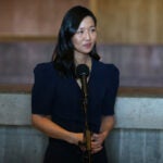 Mayor Michelle Wu wears a dark colored dress and stands at a microphone at a press conference inside Boston City Hall.