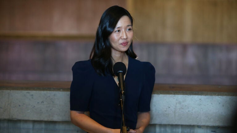 Mayor Michelle Wu wears a dark colored dress and stands at a microphone at a press conference inside Boston City Hall.