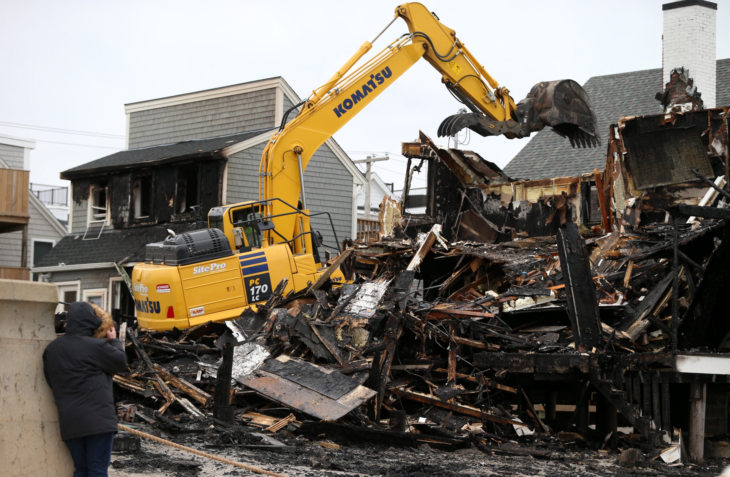 A piece of construction machinery works to demolish the charred remains of a Scituate home destroyed in a five-alarm fire Friday night. It is daytime, and a person in a heavy winter coat is seen in the foreground, facing the demolition with their back to the camera.