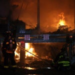A firefighter in gear looks on as flames continue to burn among the remains of several waterfront homes. Though it is nighttime, the flames illuminate the scene in a dim orange light.