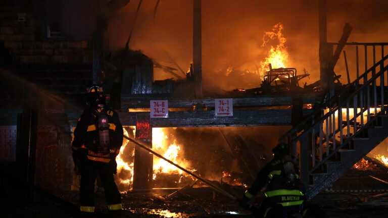 A firefighter in gear looks on as flames continue to burn among the remains of several waterfront homes. Though it is nighttime, the flames illuminate the scene in a dim orange light.