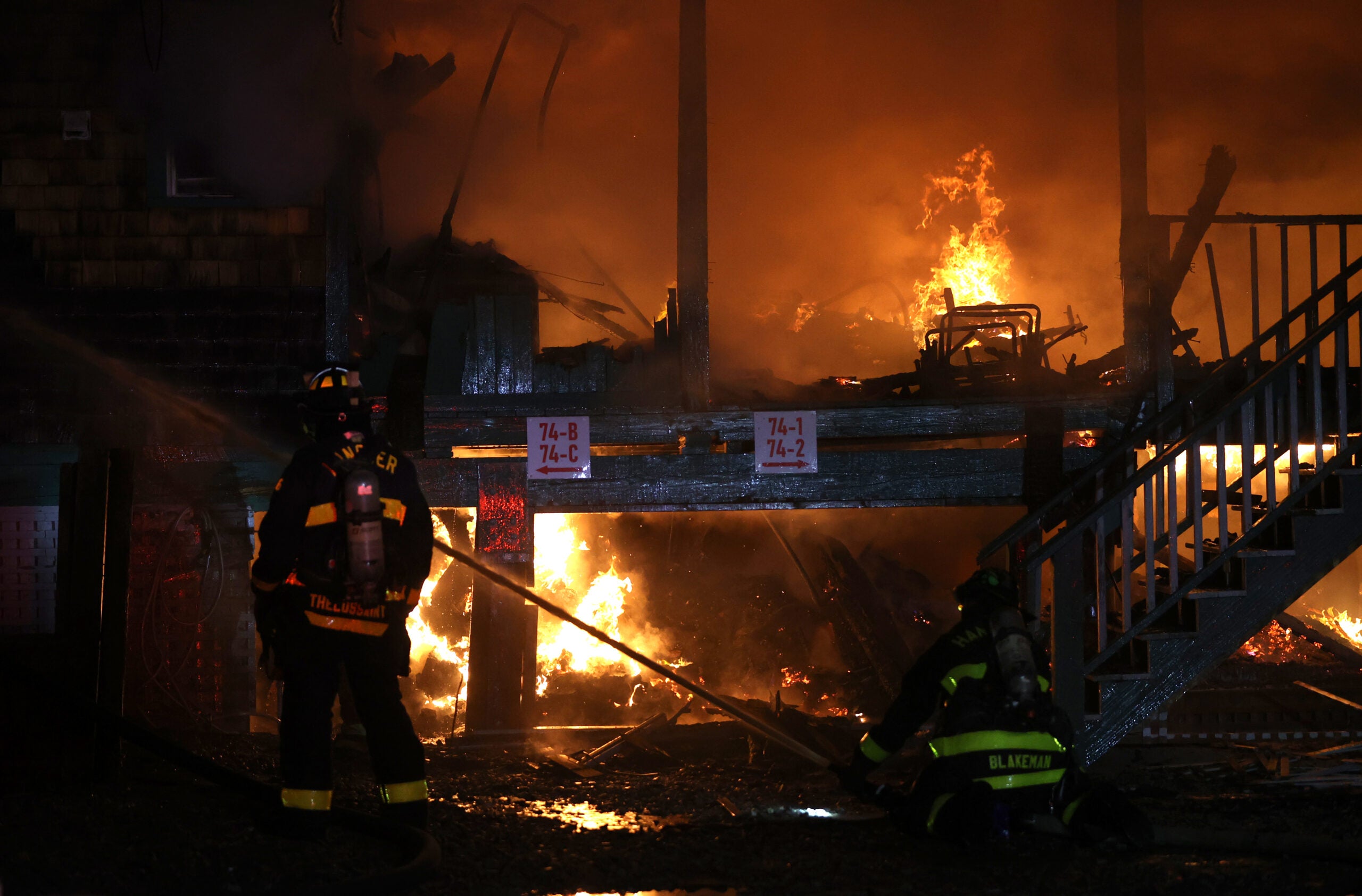 A firefighter in gear looks on as flames continue to burn among the remains of several waterfront homes. Though it is nighttime, the flames illuminate the scene in a dim orange light.