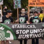 alt = Demonstrators, wearing black Starbucks union shirts and carrying colorful signs reading things like "Starbucks, stop union busting," protest outside a closed Starbucks location last year in Seattle.