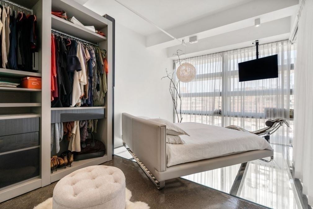 A bedroom with doorless closets, a bed without a footboard, a wall of windows, and a glass floor.