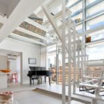 A room with a wall of windows, a grand piano, tile flooring, and a staircase with glass treads.