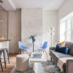 A room with griege walls, brick walls painted white, wood flooring, a kitchen peninsula with seating, and modern furniture that includes a white pouf, blue Eames chairs, and a gray couch with exposed legs and straight lines.