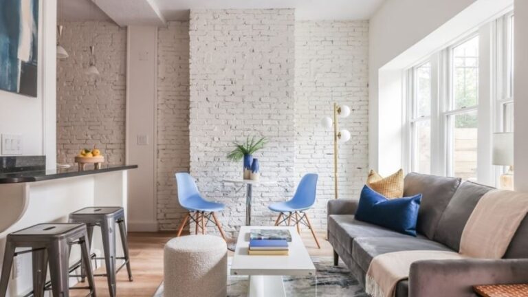 A room with griege walls, brick walls painted white, wood flooring, a kitchen peninsula with seating, and modern furniture that includes a white pouf, blue Eames chairs, and a gray couch with exposed legs and straight lines.