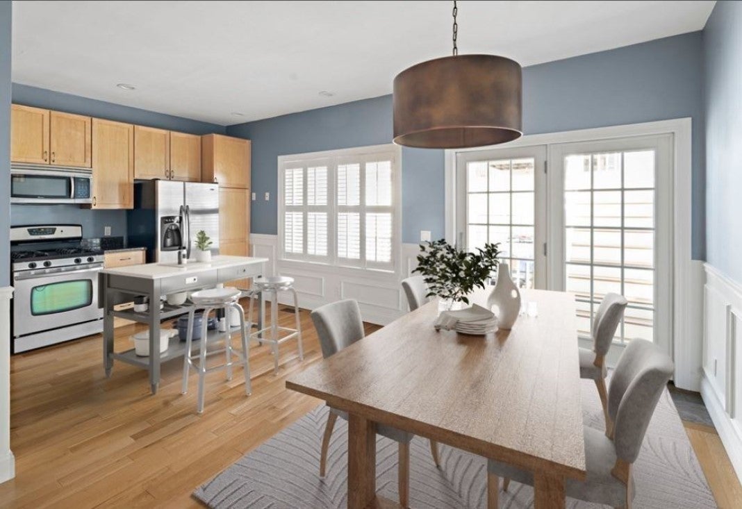 A view into a kitchen/dining area in an East Boston home with a wooden table, a copper drum shade light, wood cabinetry, and a metal island with seating for two.