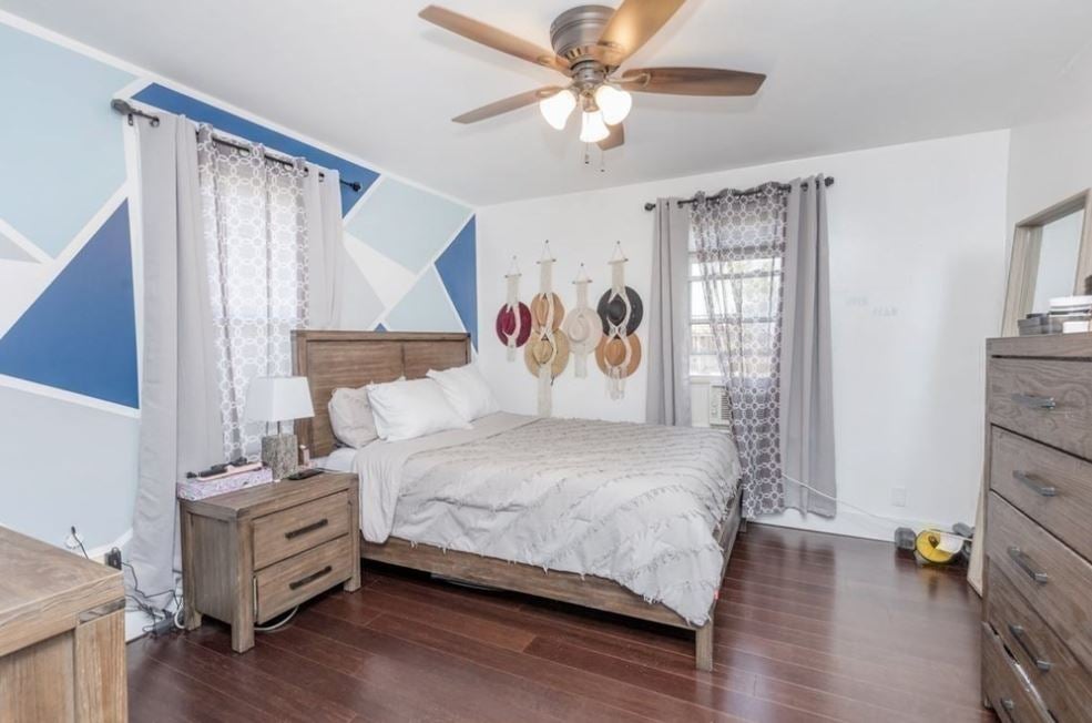 A bedroom with hardwood flooring, a ceiling fan, a bed with only a headboard, a matching light-colored wood, two-drawer night stand, two windows with sheer curtains, and an accent wall comprised of triangles in various shades of blue.