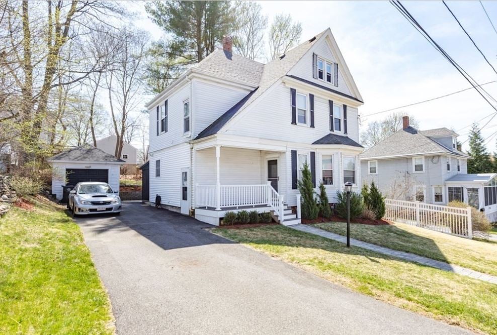 A white Colonial with sharply angled rooflines, a detached garage, a small front porch off to the side, and a green lawn the driveway bisects.