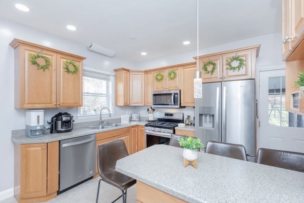 A kitchen with light-colored wood cabinetry, stainless steel appliances, wreaths on the upper cabinets, an island with a gray countertop, and stools with backs.