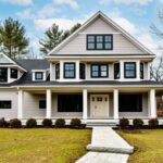 A Victorian with gray shingles and clapboard, a farmer's porch with white columns, no shutters, and black windows. A paving stone walkway leads up to the home, which is fronted by immature shrubs. The grass is green but the deciduous trees are bare.