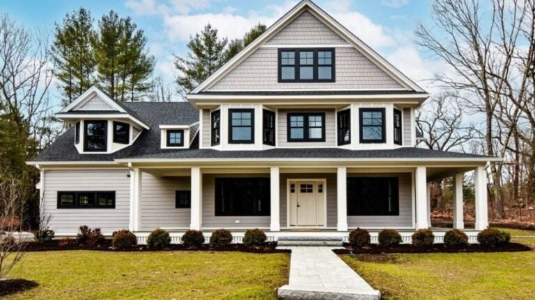 A Victorian with gray shingles and clapboard, a farmer's porch with white columns, no shutters, and black windows. A paving stone walkway leads up to the home, which is fronted by immature shrubs. The grass is green but the deciduous trees are bare.