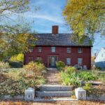 A brick-red Ipswich Colonial with a cedar shake room, no shutters, a stone walkway with mature plantings, and nine windows on the front facade.