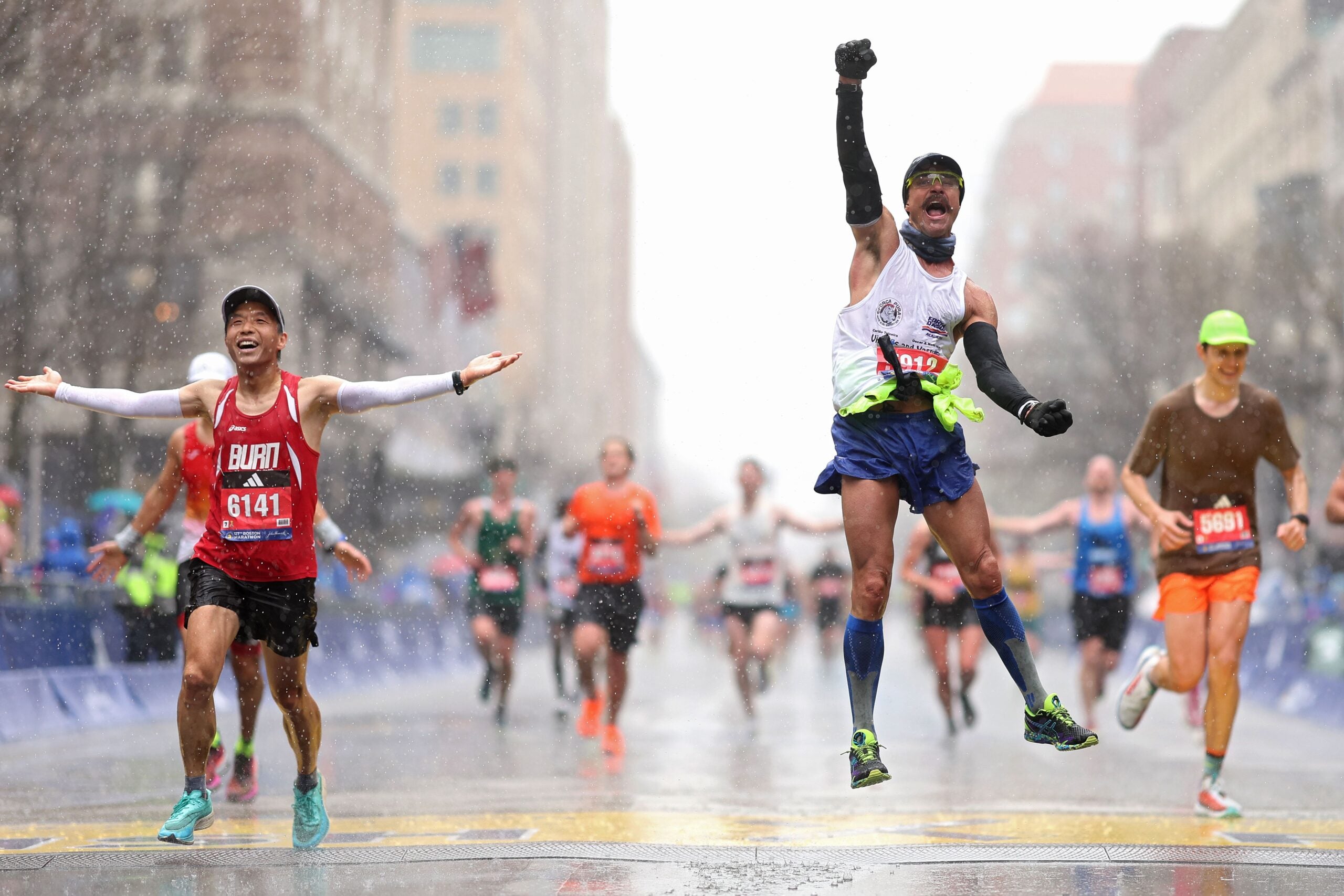 Fernando Ferreira celebrates in the rain as he crosses the finish line next to Zhenfei Lu, left, during the 127th Boston Marathon on April 17, 2023 in Boston, Massachusetts. 