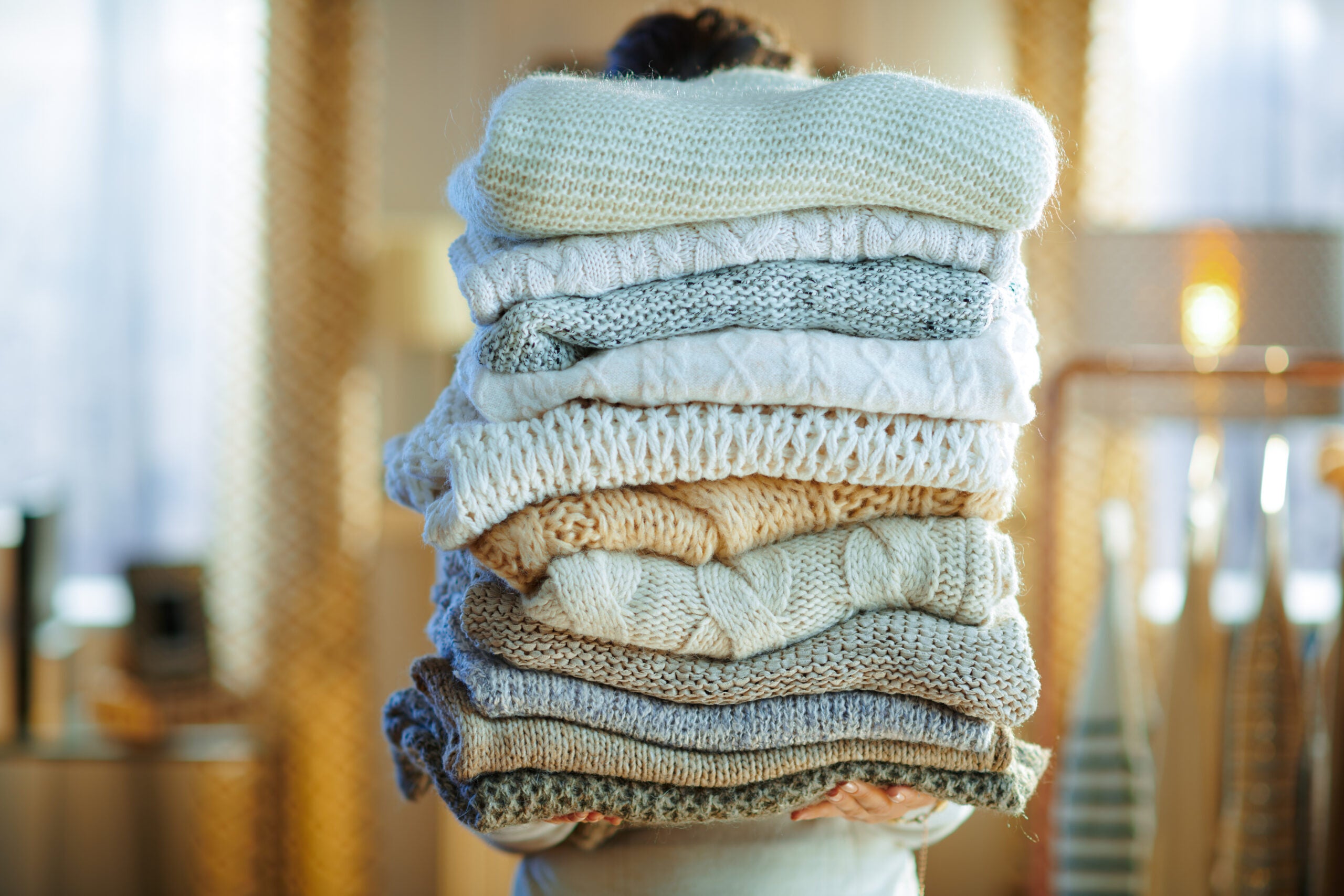 stylish woman in white sweater and skirt in the modern house in sunny winter day holding huge pile of sweaters.