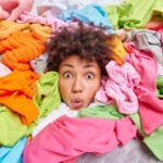 apartment storage: Decluttering second hand spring cleaning fast fashion and organization of life. Stunned Afro American woman with curly hair looks through big heap of colorful clothes puts things in order at closet