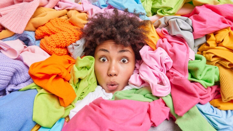apartment storage: Decluttering second hand spring cleaning fast fashion and organization of life. Stunned Afro American woman with curly hair looks through big heap of colorful clothes puts things in order at closet