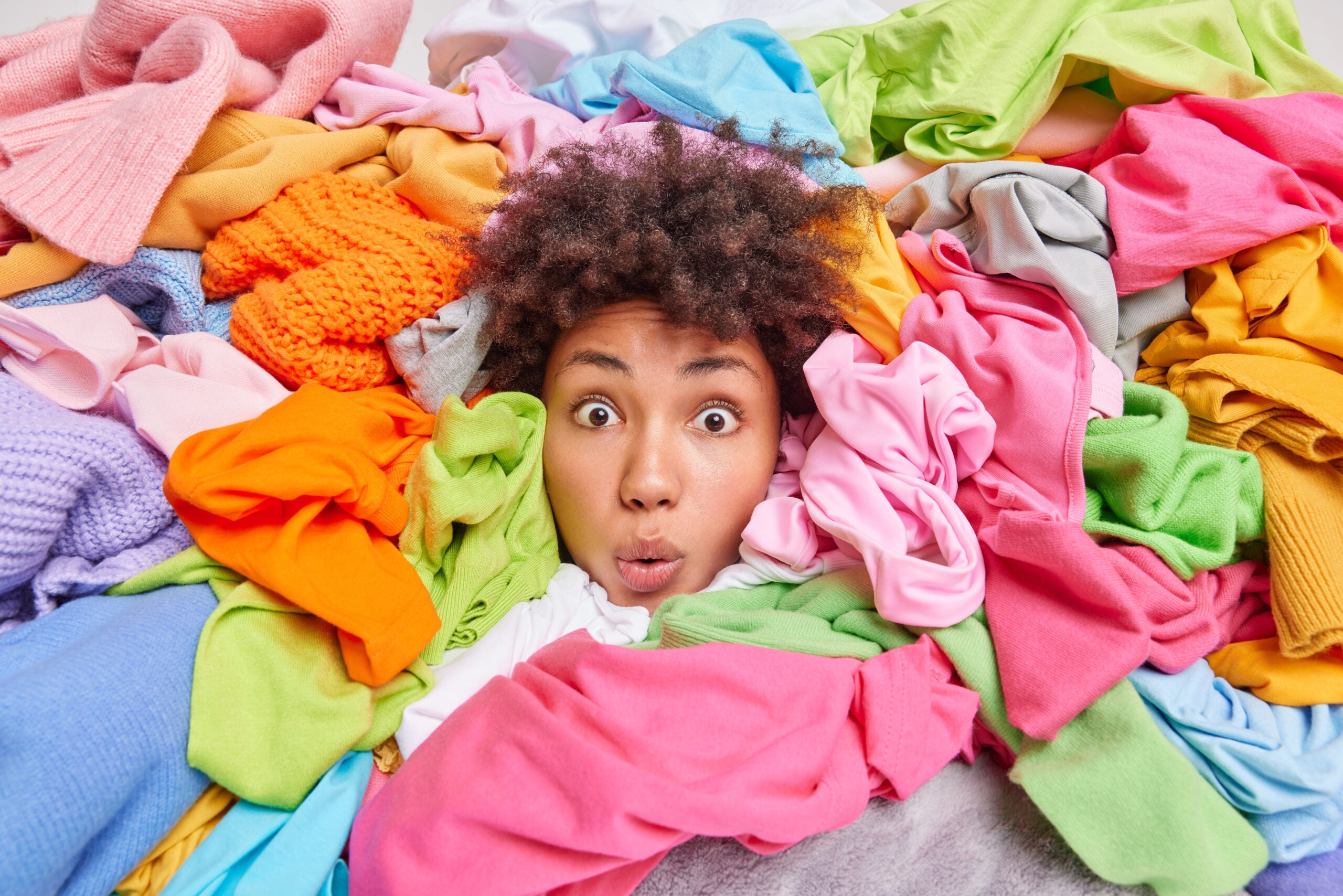 apartment storage: Decluttering second hand spring cleaning fast fashion and organization of life. Stunned Afro American woman with curly hair looks through big heap of colorful clothes puts things in order at closet