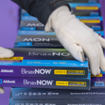 Gloved hands hold boxes of at-home COVID tests. The background is tinted purple.