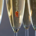 A small scuba diver figure in a red suit, black helmet, and flippers floats in a flute of champagne, photographed as a close-up.