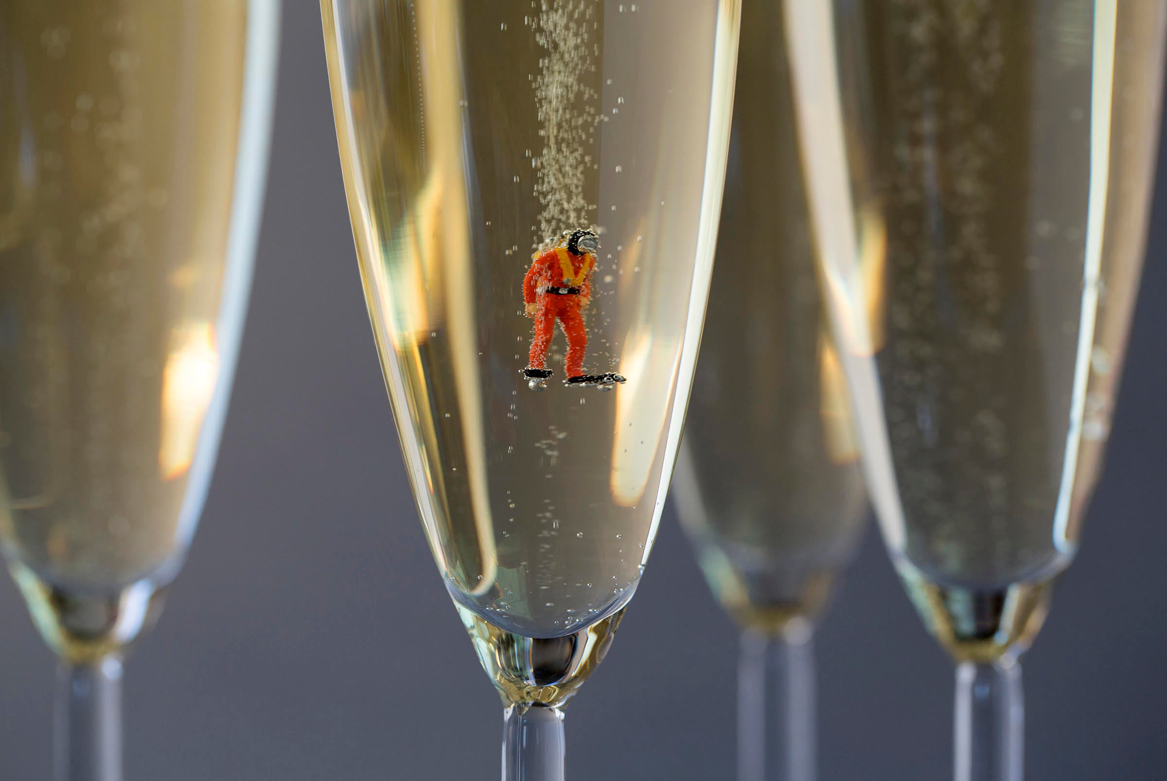 A small scuba diver figure in a red suit, black helmet, and flippers floats in a flute of champagne, photographed as a close-up.