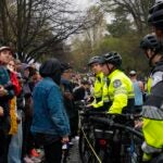 Marathon spectators are shown dressed for wet and chilly weather on one side of a rope barrier. On the other side, several police officers in fluorescent yellow jackets and helmets stand holding bicycles, blocking the spectators from the marathon course.