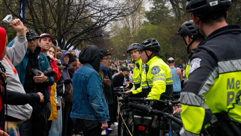 Marathon spectators are shown dressed for wet and chilly weather on one side of a rope barrier. On the other side, several police officers in fluorescent yellow jackets and helmets stand holding bicycles, blocking the spectators from the marathon course.