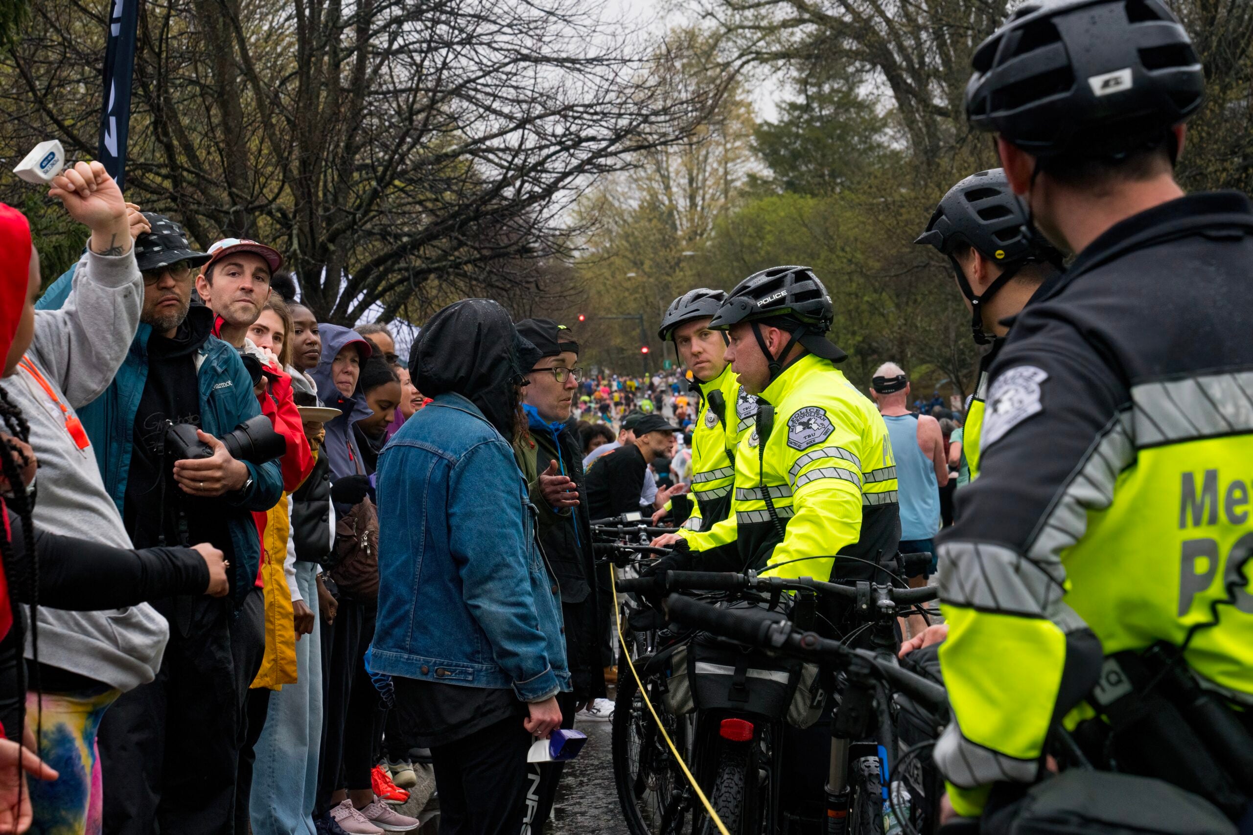 Marathon spectators are shown dressed for wet and chilly weather on one side of a rope barrier. On the other side, several police officers in fluorescent yellow jackets and helmets stand holding bicycles, blocking the spectators from the marathon course.
