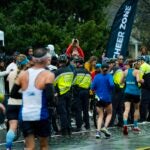 A line of police officers in fluorescent yellow and black jackets stands between a crowd of Boston Marathon spectators and the race course, their backs to the runners in the foreground.