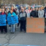 Dozens of people stand outdoors during a standout protest, some holding cardboard and paper signs in support for Easthampton superintendent finalist Dr. Vito Perrone.