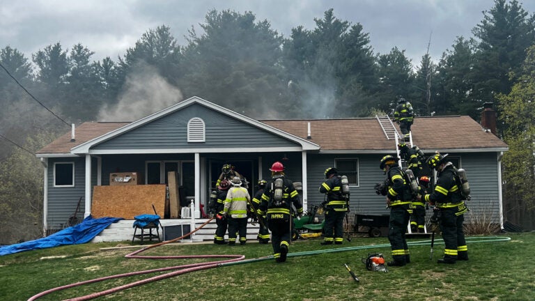 A group of firefighters surround a smoking blue house working to put out a fire.
