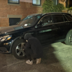 A man in a black hoodie deflates the tire of a black SUV