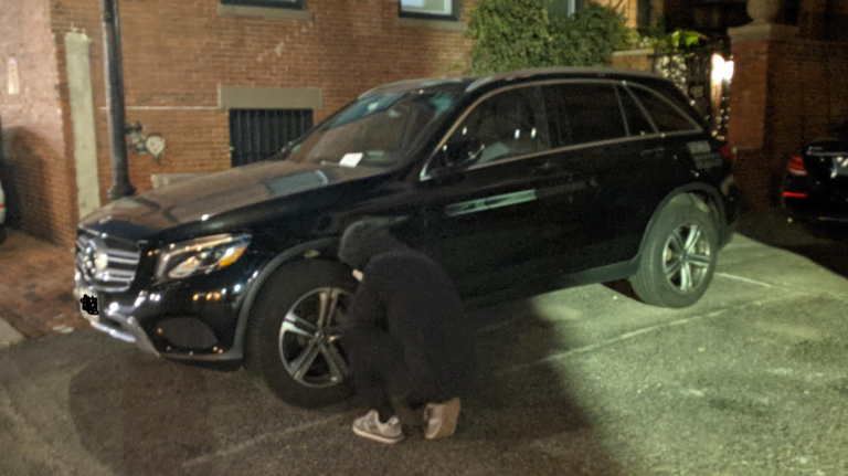 A man in a black hoodie deflates the tire of a black SUV