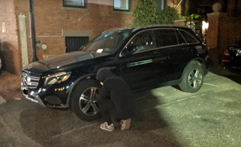 A man in a black hoodie deflates the tire of a black SUV