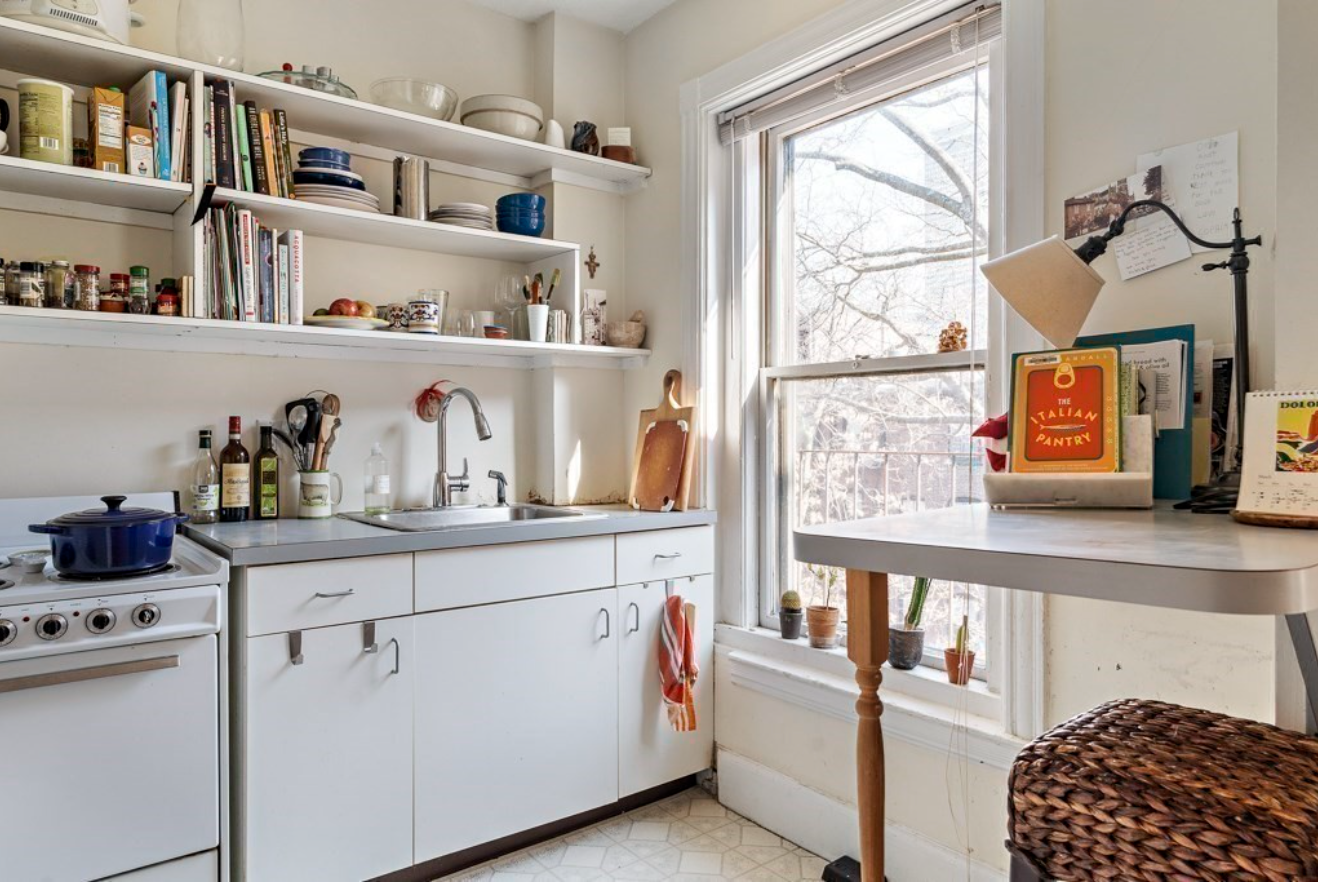 The kitchen has a single-hung window, floating shelves, and cream-colored walls.