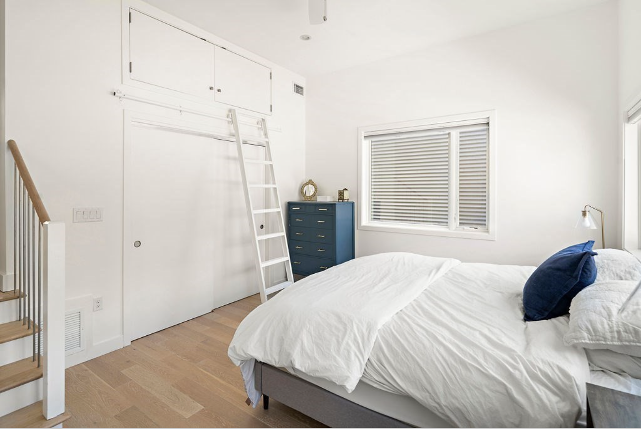 A bedroom with white walls and hardwood floors. A sliding ladder leads to overhead storage.