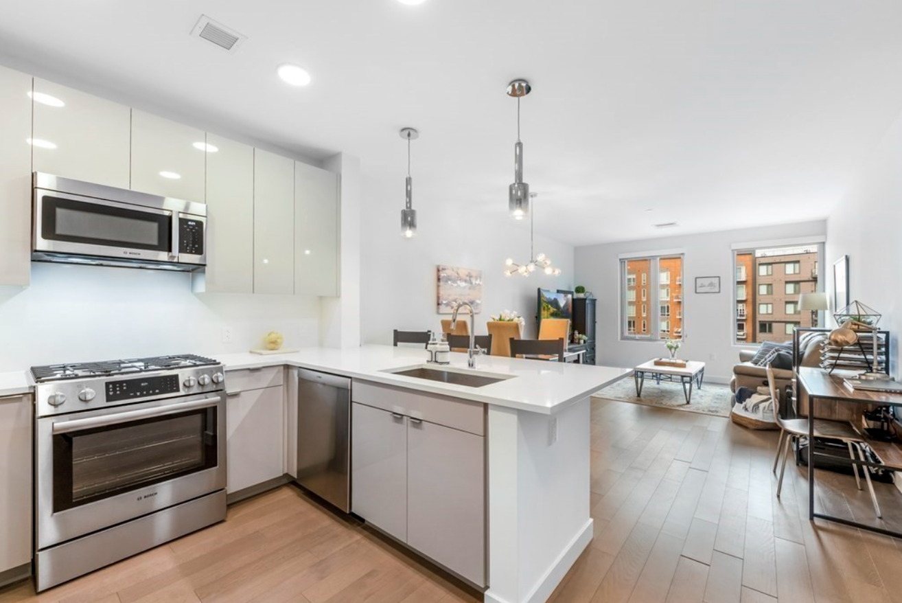 The kitchen has white flat-panel cabinets and stainless steel appliances. The open floor plan leads to the living room, with white walls and two single-hung windows.