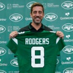 Aaron Rodgers holding up his new Jets jersey with the number 8 on it.