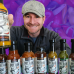 A man sits behind a row of hot sauce bottles and holds one up for the camera. The background is a purple-tinted picture of hot peppers.