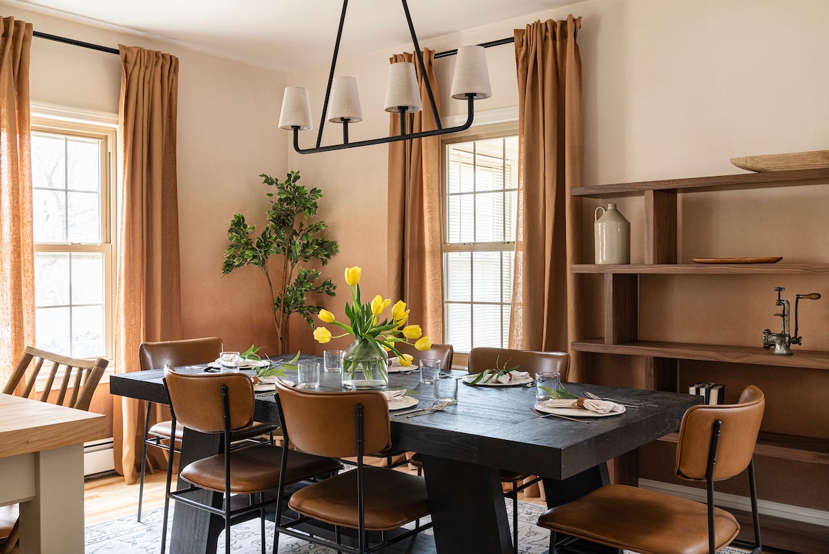 A light-colored dining room with tan draperies, a black table, modern chairs upholstered in a tan fabric, and a bookshelf.