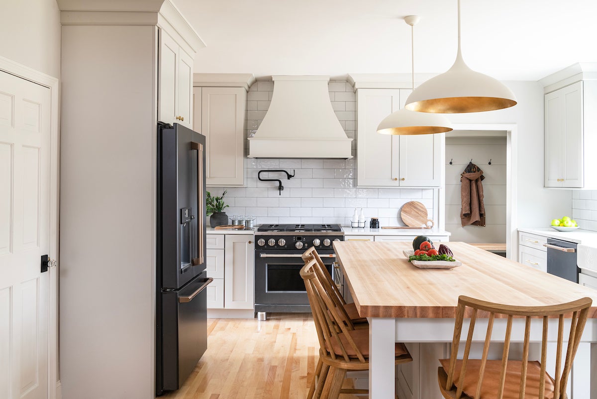 A kitchen with white cabinetry, a long table with wooden, arched spindle chairs, and white round-domed pendant lights with a bronze finish inside.