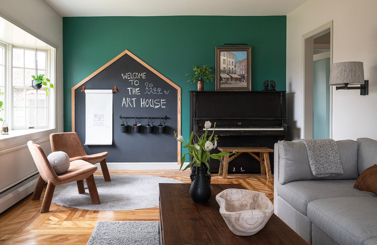 A room with a green accent wall, hardwood flooring, gray area rugs, a wood coffee table, a gray couch, a black piano, with a wood bench, two wooden chairs, and a house-shaped chalkboard that says 