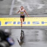 Emma Bates crosses the Boston Marathon's yellow finish line with her arms outstretched.