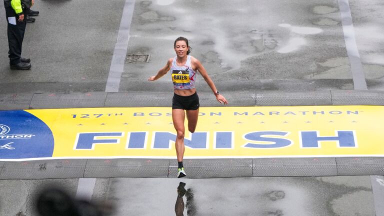 Emma Bates crosses the Boston Marathon's yellow finish line with her arms outstretched.