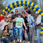 Gates and his planning committee smile and give thumbs-up alongside the last marathon runner of 2022. The group is under a yellow-and-blue balloon arch.