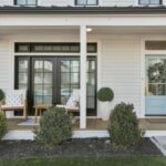 An open house is being held at a home with a short front porch with white columns and trim, a wood floor. Two white chairs are set before black French doors. The front door is robin's egg blue with four glass panes. Immature bushes are lined up in front of the porch.