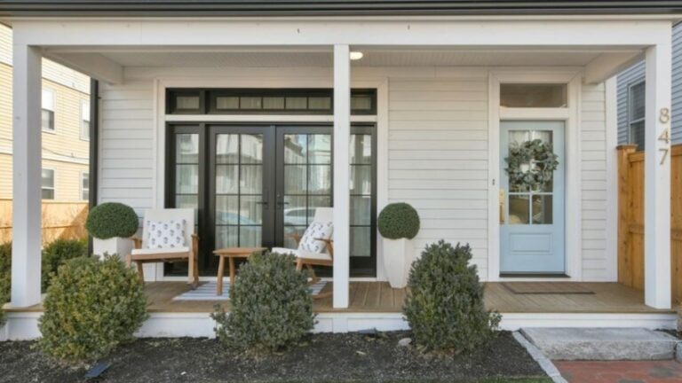An open house is being held at a home with a short front porch with white columns and trim, a wood floor. Two white chairs are set before black French doors. The front door is robin's egg blue with four glass panes. Immature bushes are lined up in front of the porch.